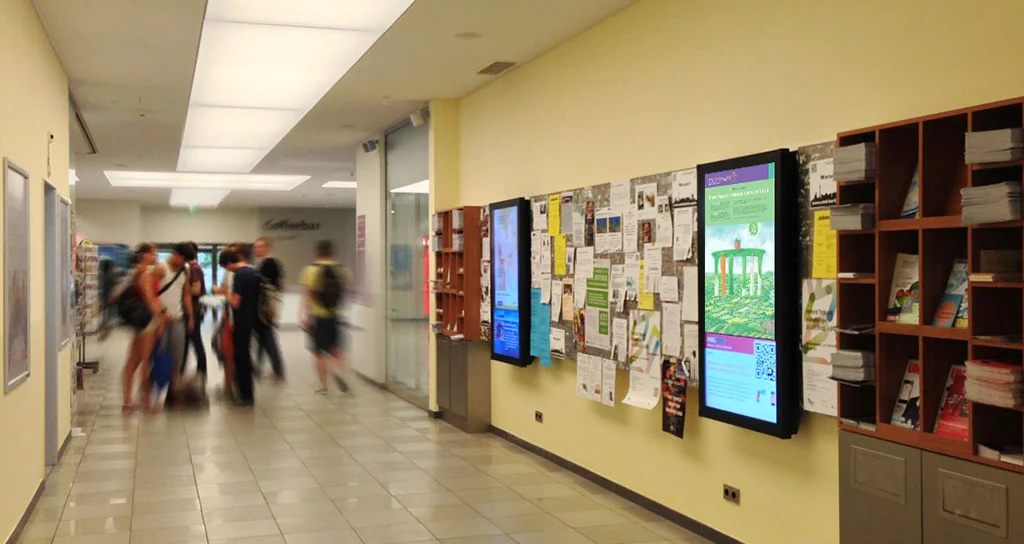 Digitale Bildschirme in einem Hochschulgebäude, an denen Studierende vorbeigehen
